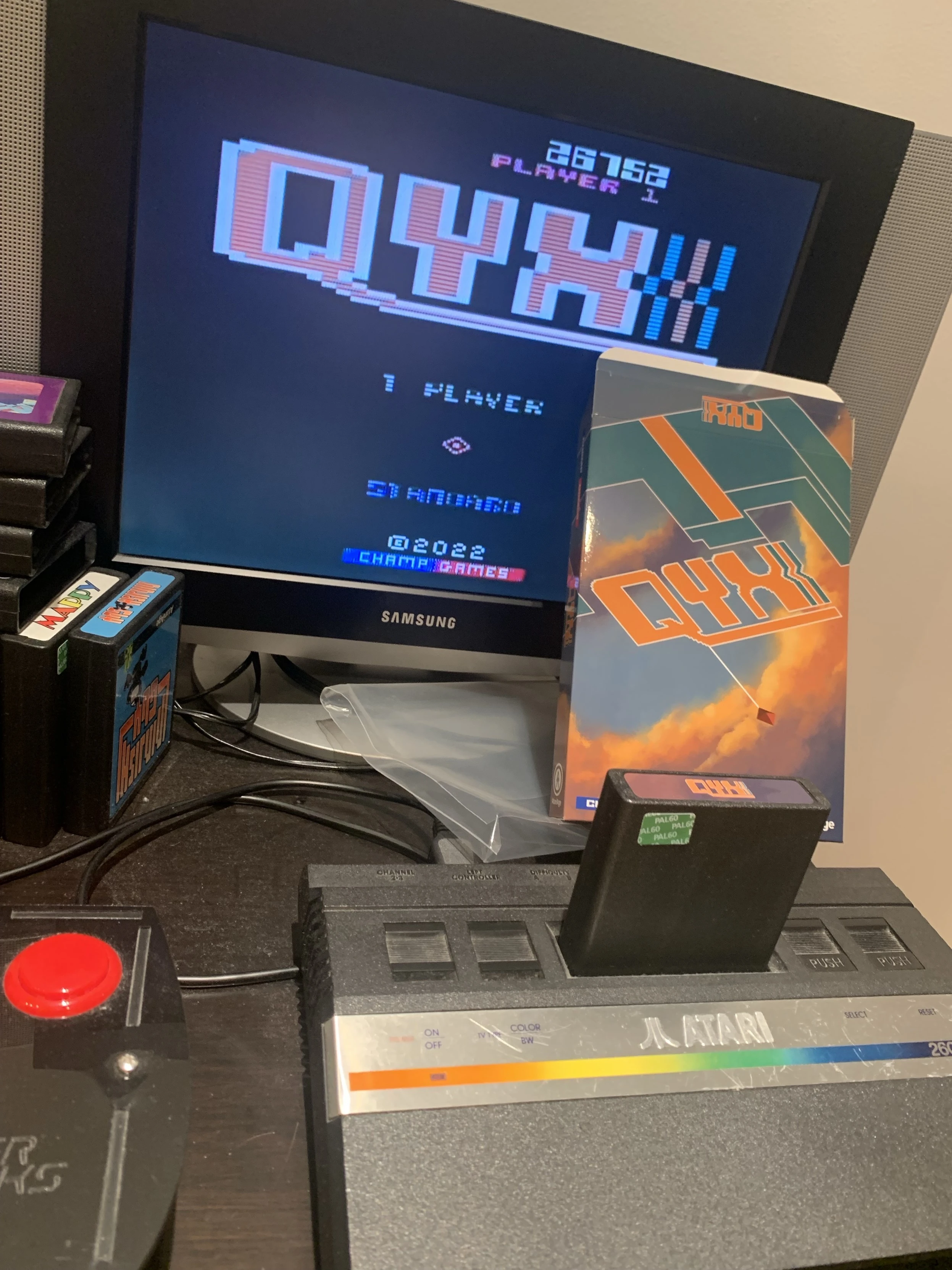 An Atari console is displayed on a table, with a game cartridge labeled "QYX" inserted. The screen shows the game title and score. A game box for "QYX" is also visible, alongside other game cartridges.