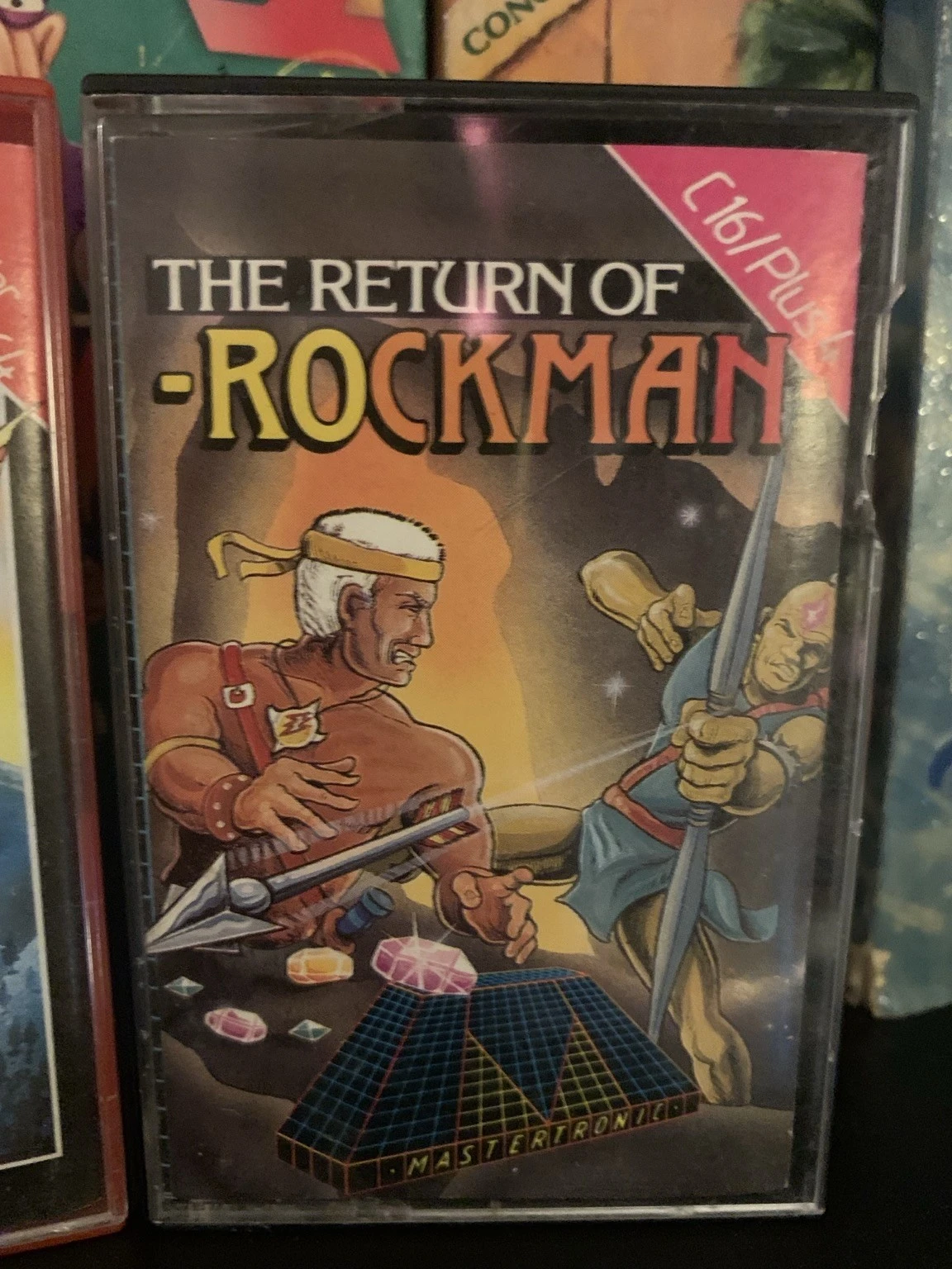 Image of a cassette cover for a video game titled "The Return of Rockman." It features two characters in combat, one muscular with a spear and the other in blue clothing wielding a sword. Below them, a grid design with colorful gems and the Mastertronic logo below. It is for the Commodore C16 and Plus4 line of machines. 