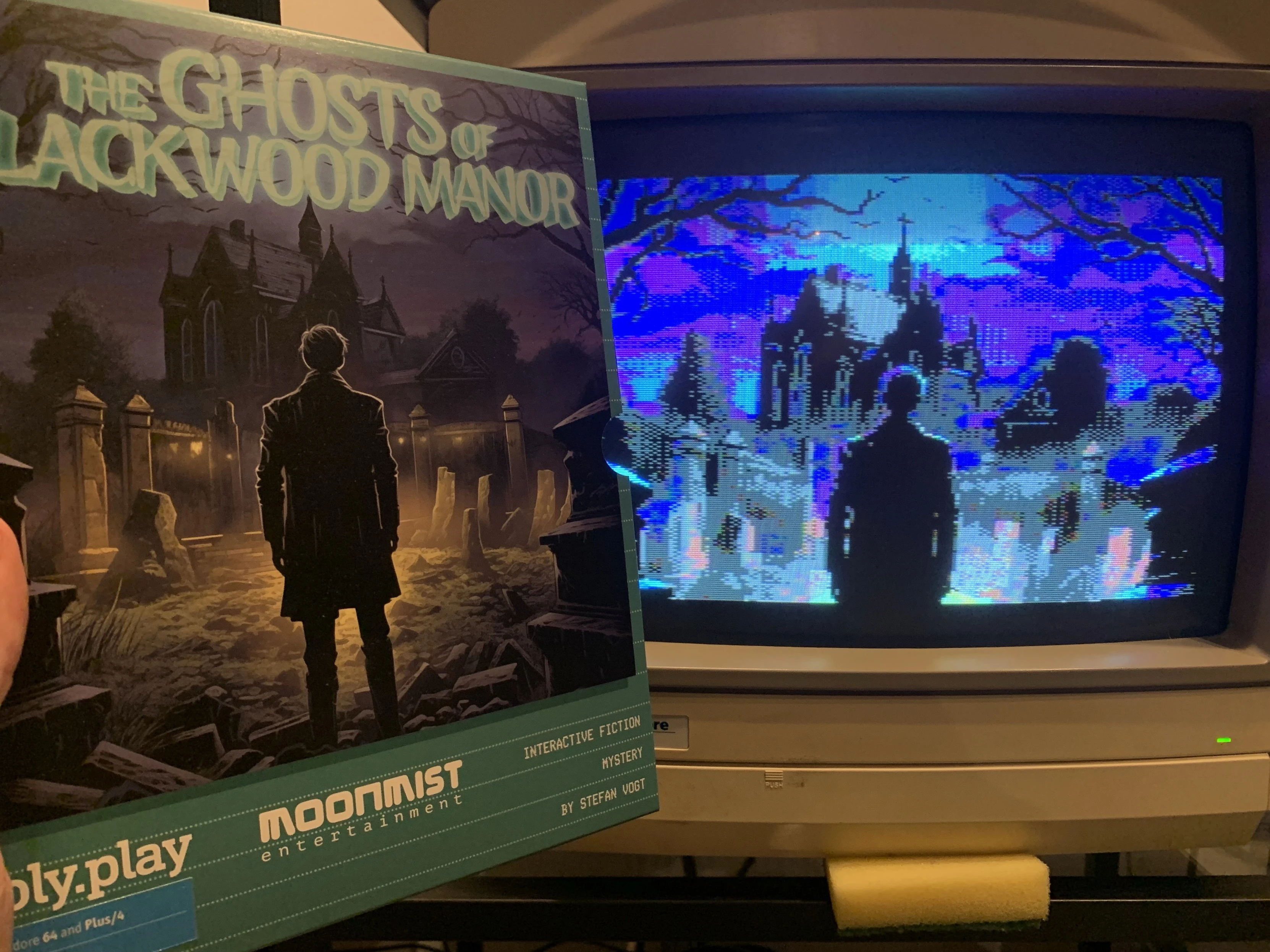 An individual is holding a box for the game "The Ghosts of Blackwood Manor," featuring a dark, haunted house scene. Next to it, an old monitor displays a pixelated version of the same scene, showing a silhouette standing in front of a house. 